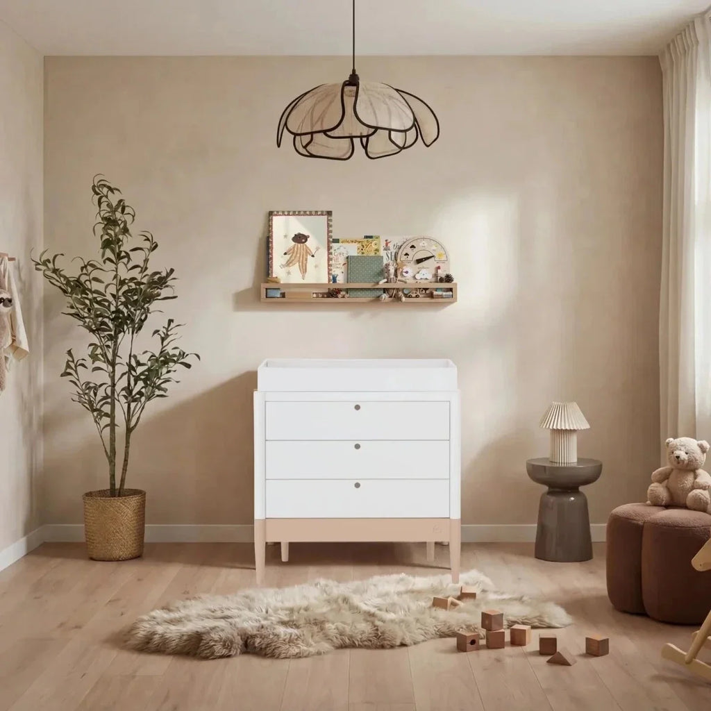 White baby dresser with three drawers, gold knobs, raised legs, powder blush bottom panel