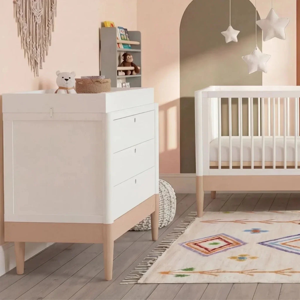 White baby dresser with drawers and powder blush base, beside white crib in nursery