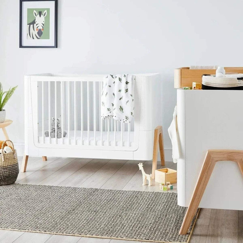 White wooden baby cot bed with natural legs and green printed blanket