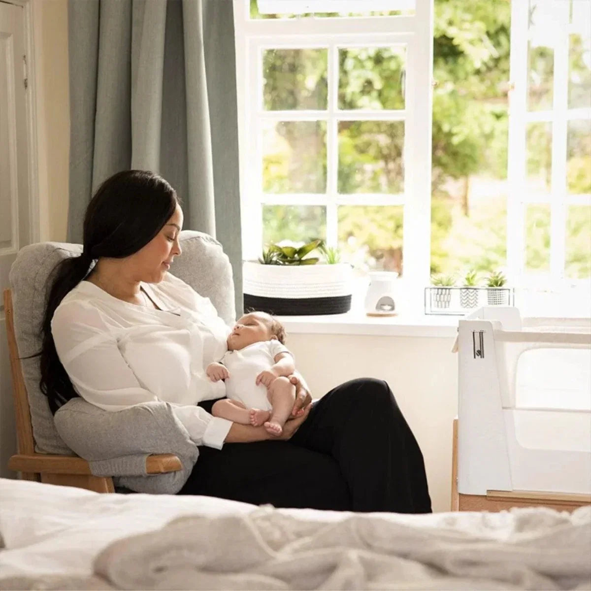 Baby seated in white bedside crib with mesh side and natural wood frame