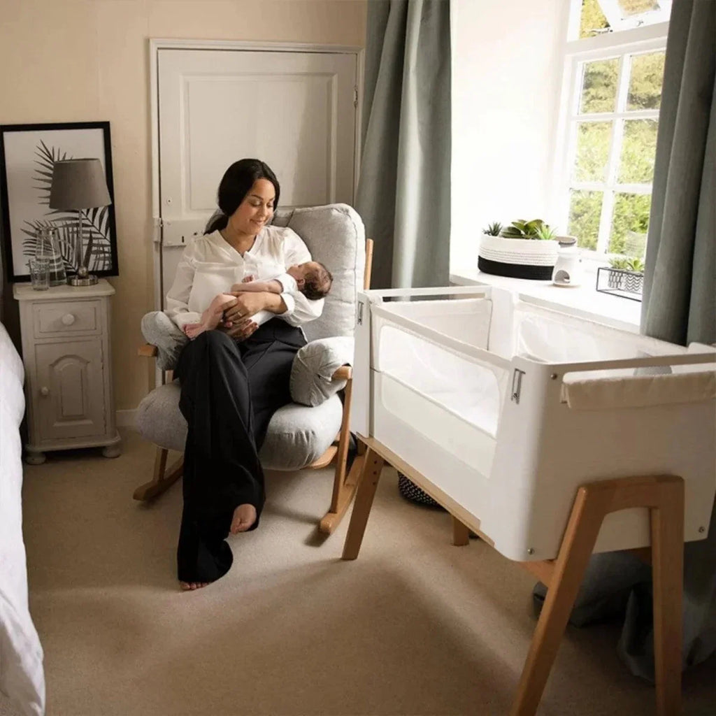 White bedside crib with open front, natural legs, and mesh bed panel