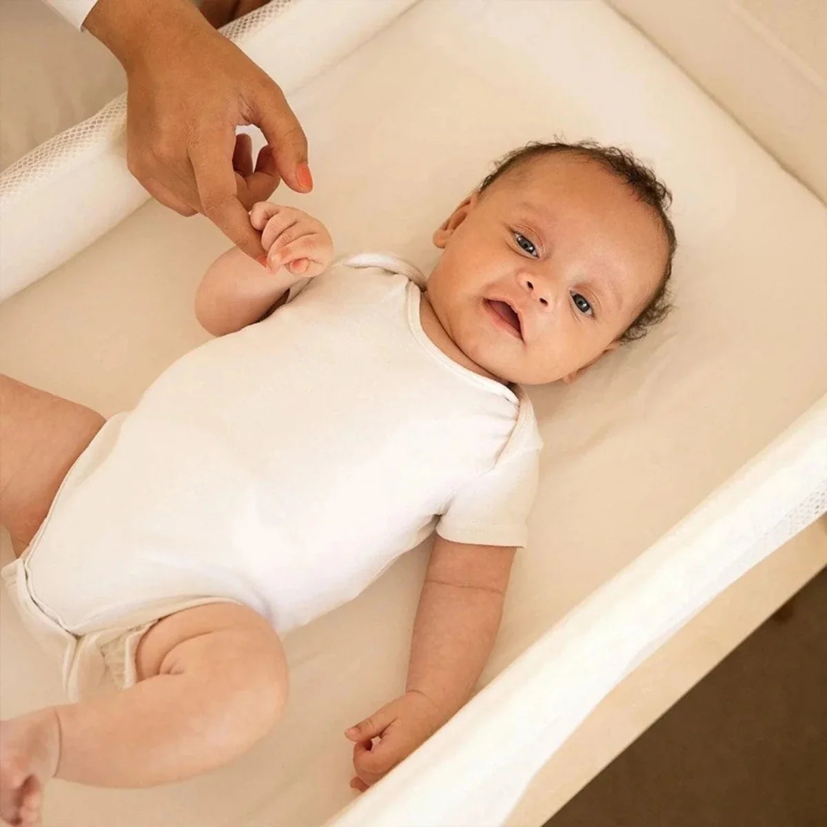 Baby lying in white bedside crib with mesh barrier and padded mattress