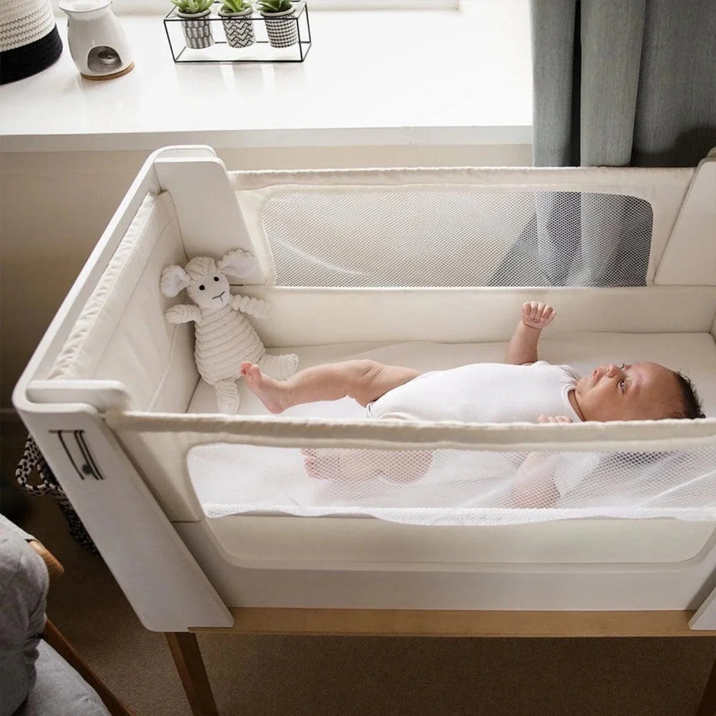 White bedside crib viewed from side with mesh guard and natural wood base