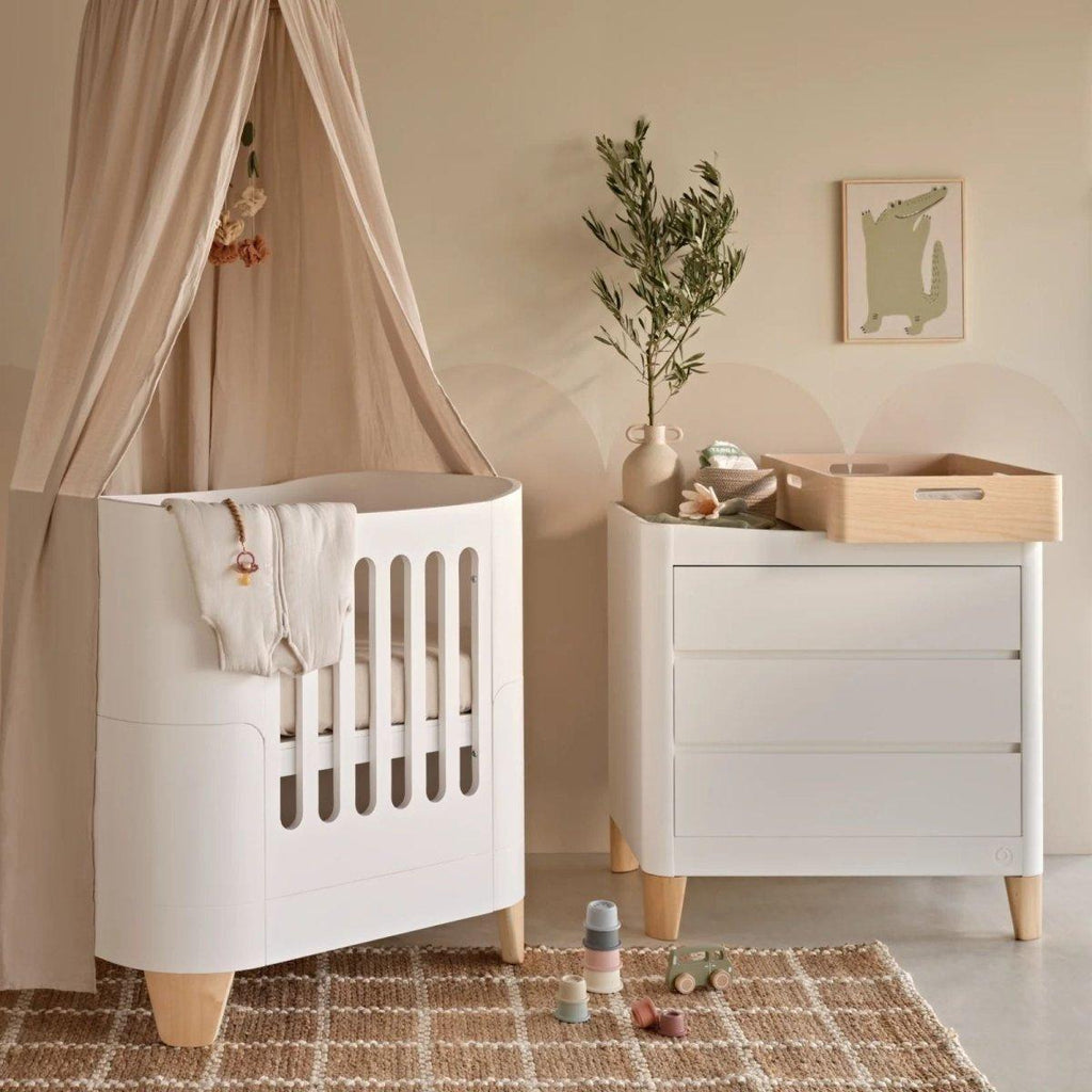 White mini cot with vertical slat sides and fitted mattress, styled in a nursery