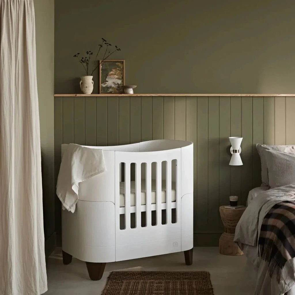 White mini cot with open slats and turned feet, shown in a nursery set