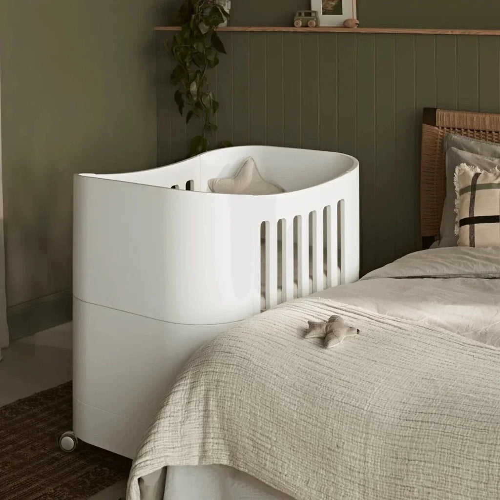 White mini cot with rounded sides and vertical safety rails, placed near a bed