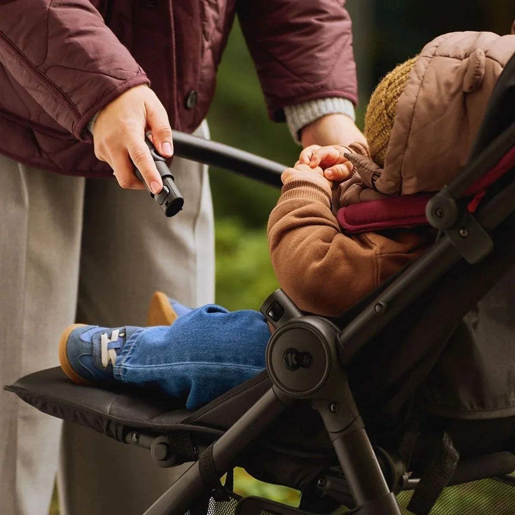 Parent adjusting stroller handle while child sits in dark ruby seat