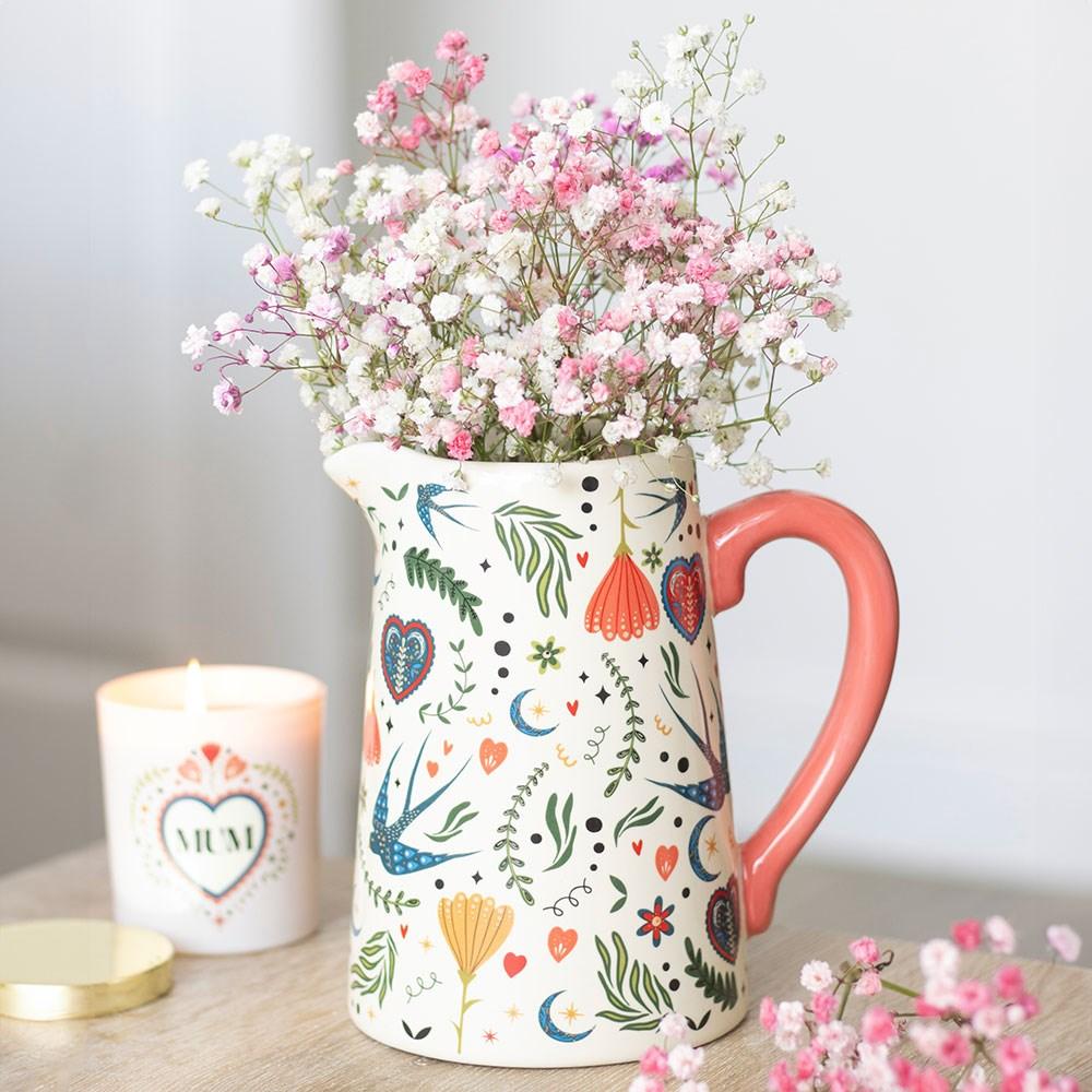 Decorative pitcher with floral patterns filled with pink and white flowers, next to a lit candle on a wooden surface.