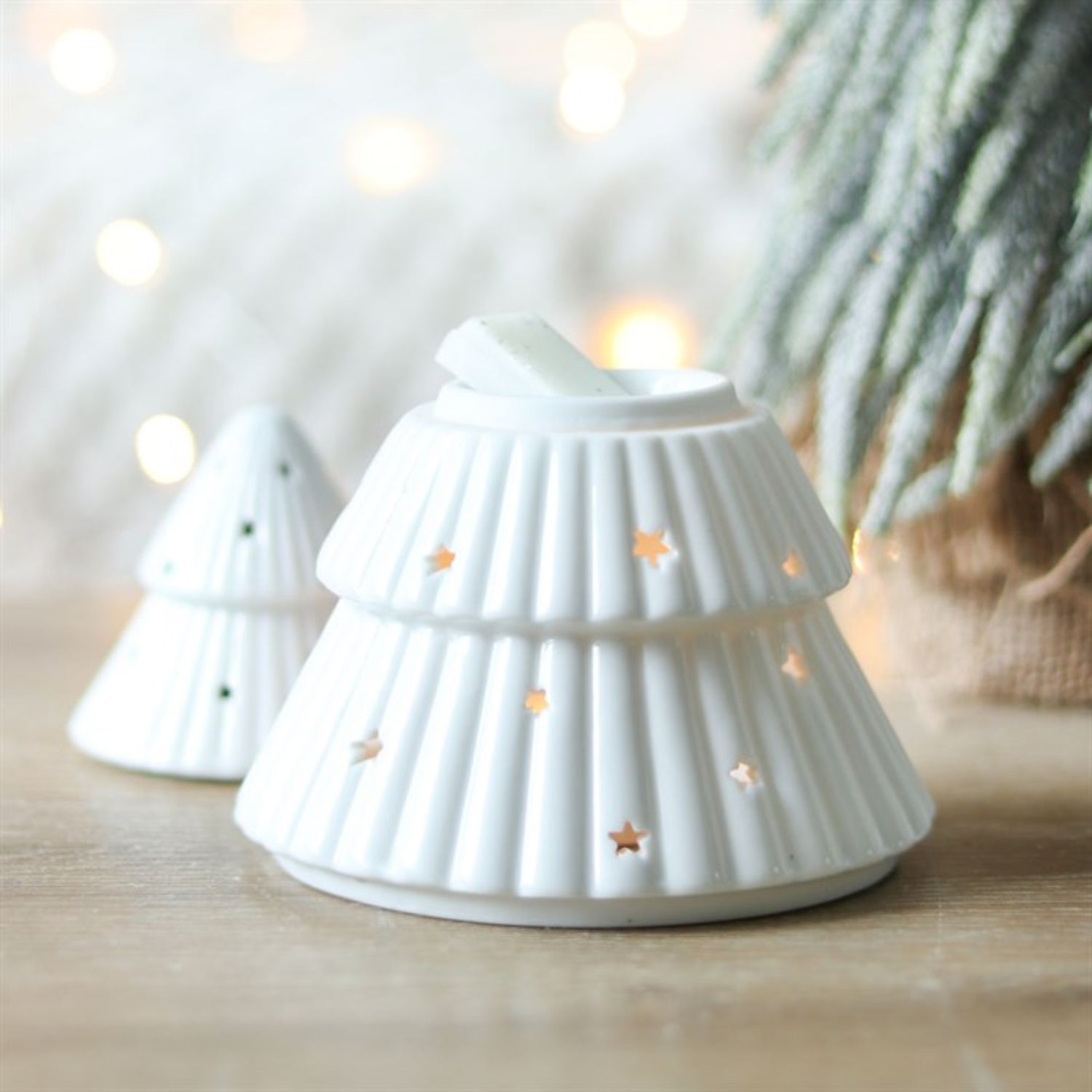 White ceramic tree-shaped candle holder with star cutouts on a wooden surface with blurred lights and plants in the background.