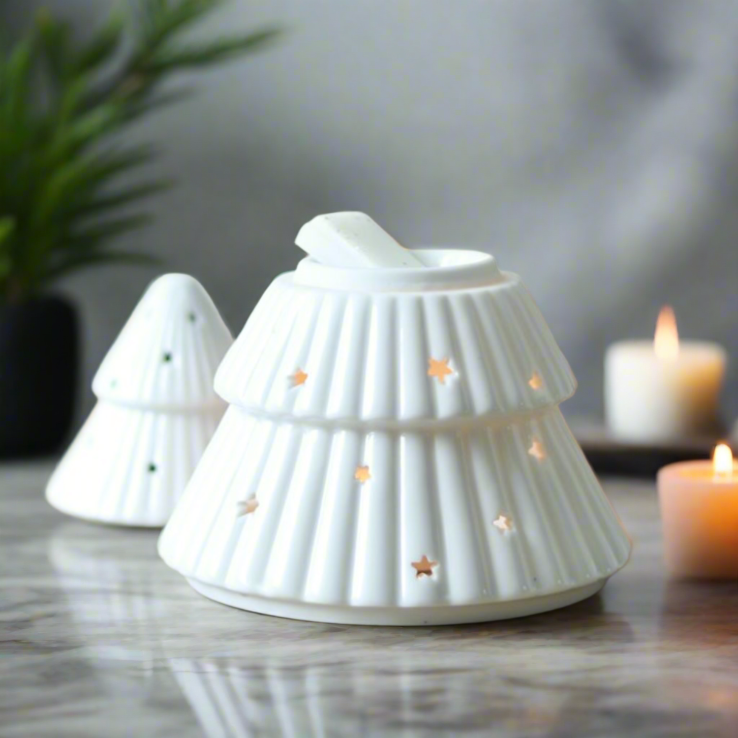White ceramic tree-shaped candle holder with star cutouts on a wooden surface with blurred lights and plants in the background.