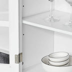 Interior shelf of the white cabinet with framed glass door, storing stacked plates and stemware