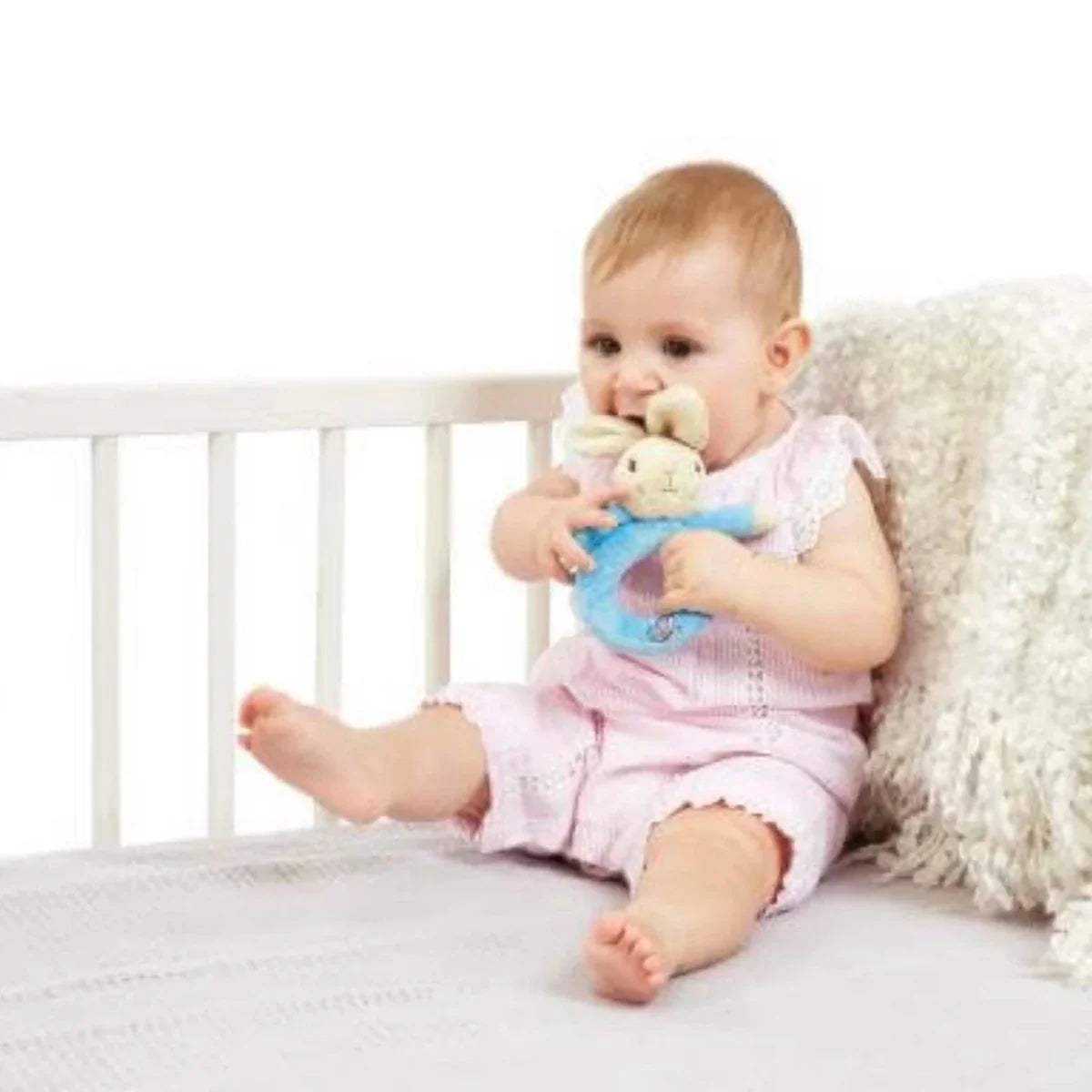Infant holding blue bunny plush ring rattle while sitting on bed
