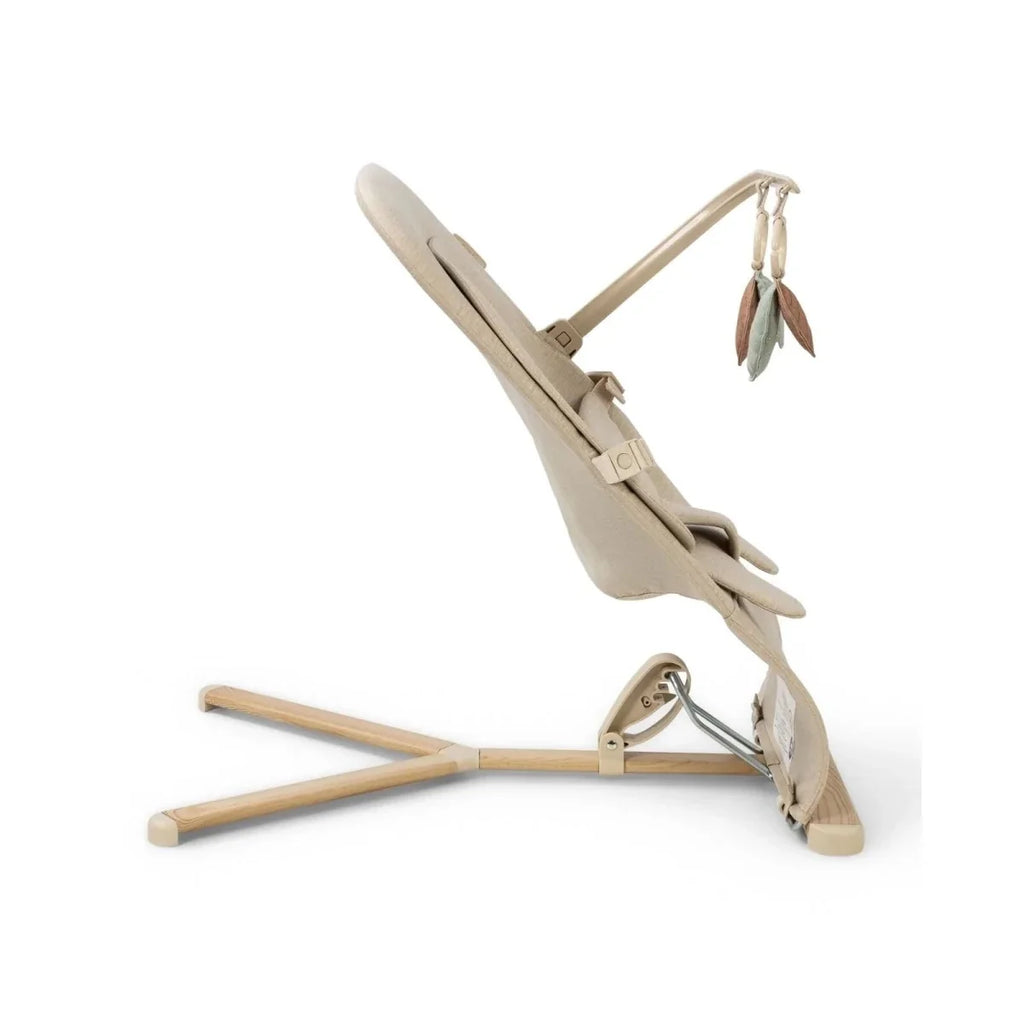Oatmeal baby bouncer in side view with reclining frame, harness straps, and leaf toy bar