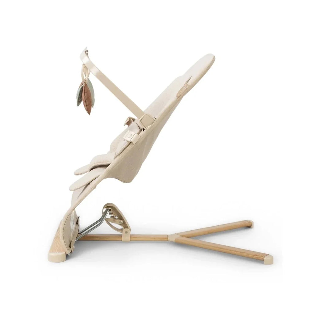 Oatmeal baby bouncer angled side view with harness straps, leaf toy arches, and wood base