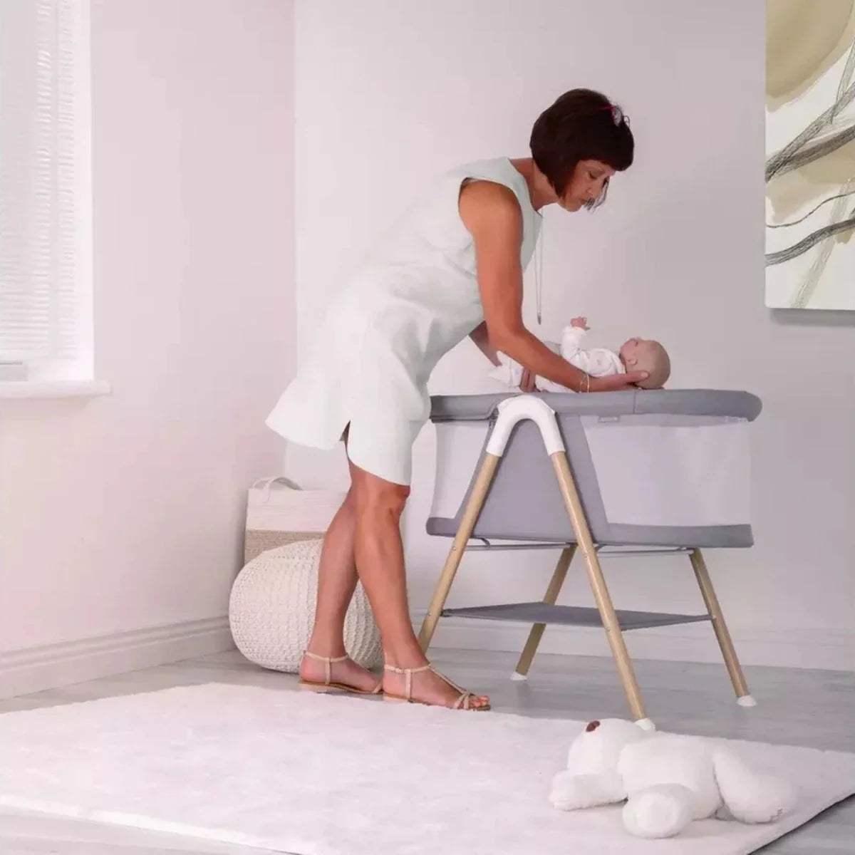 Grey snooze crib on wooden stand with caregiver leaning over baby.