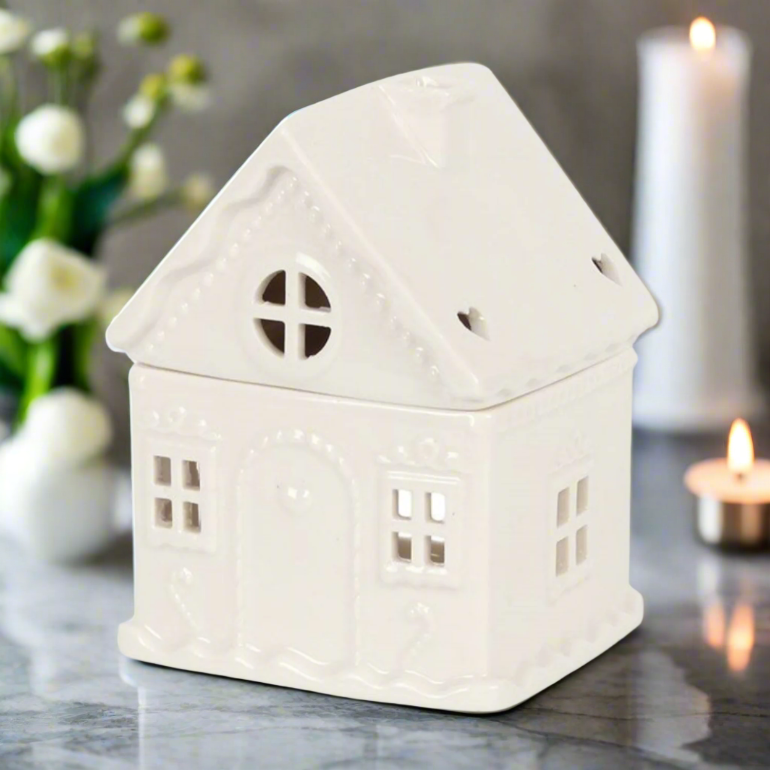 White house-shaped ceramic lantern on a marble surface with flowers and candles in the background