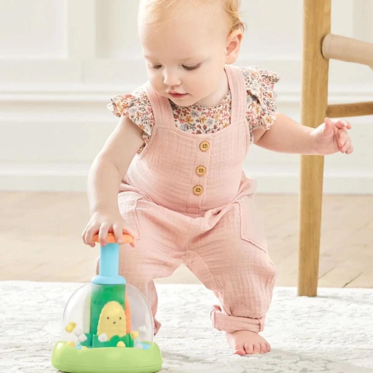 Baby in pink overalls playing with push-and-spin walker toy, green base and blue post