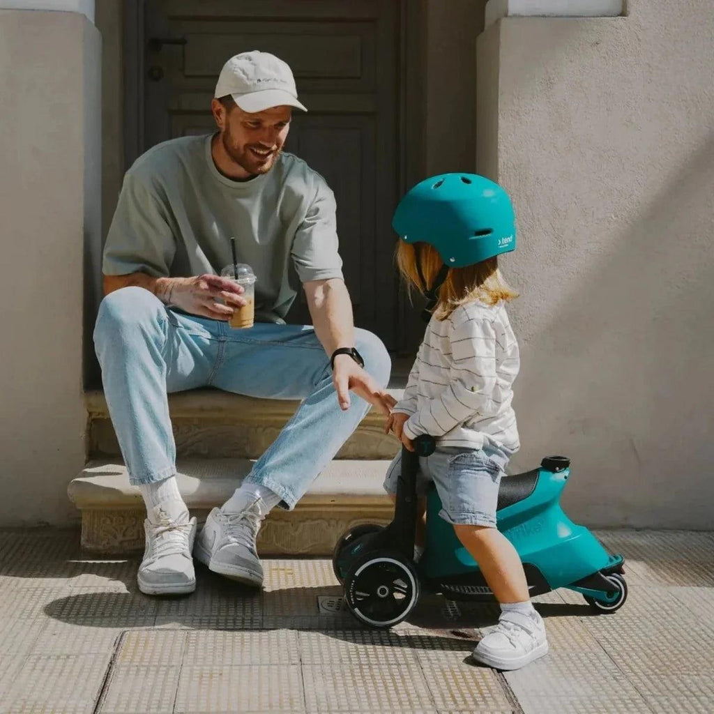 Teal ride-on scooter with child wearing a helmet, standing outdoors