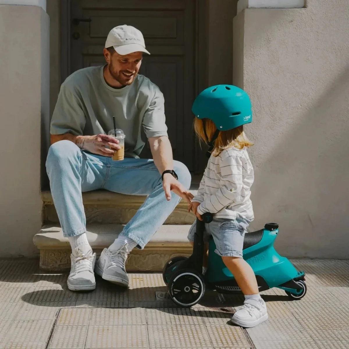 Teal ride-on scooter with child wearing a helmet, standing outdoors