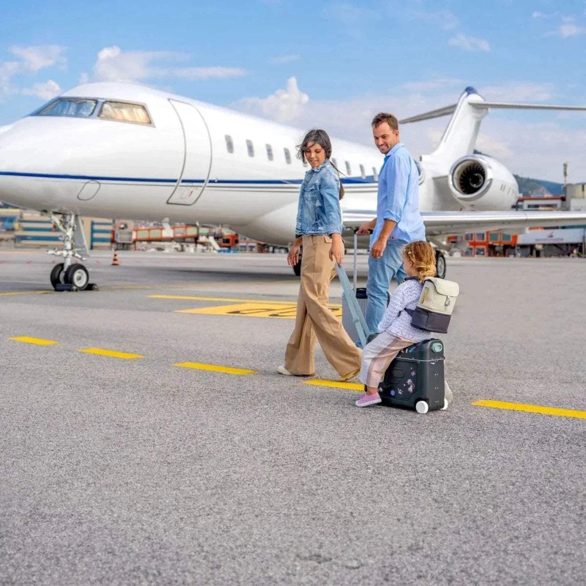 Family pulling kids ride-on suitcases at an airport beside a plane