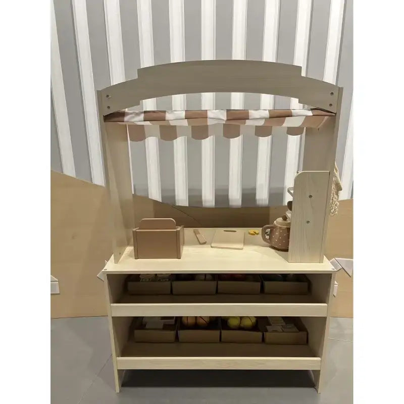 Wooden kids play shop viewed from the side with striped awning and open storage shelves