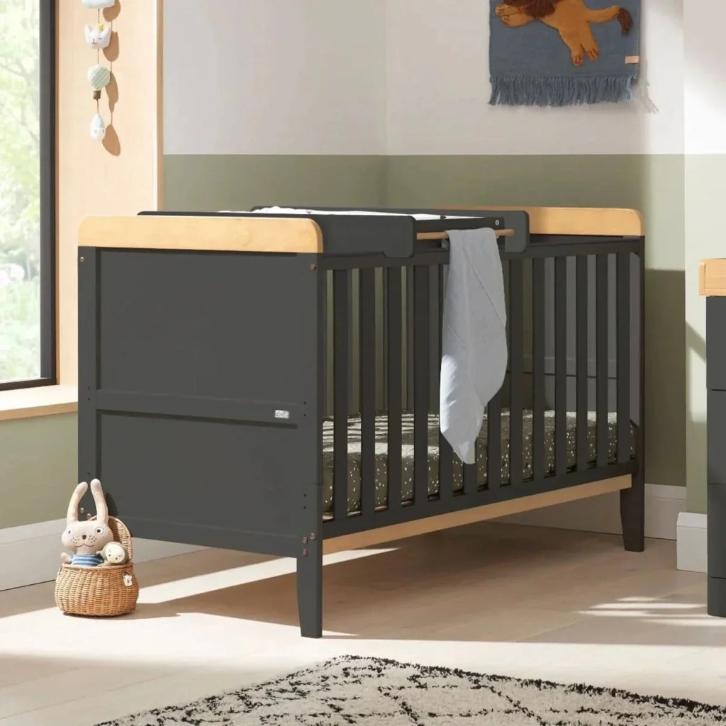 Slate cot bed with light wood rails and open side, with white cover and storage basket nearby