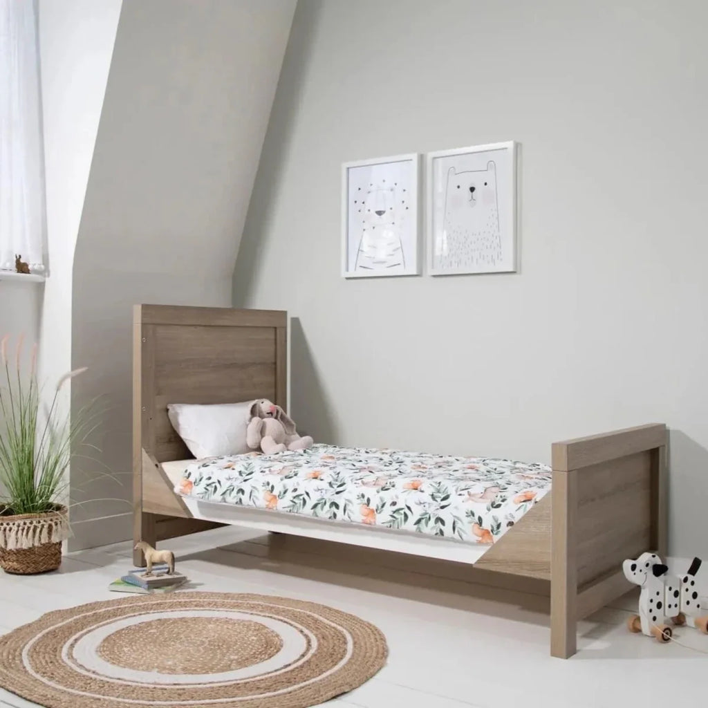 Convertible toddler bed with white slats, light oak frame, and patterned bedding