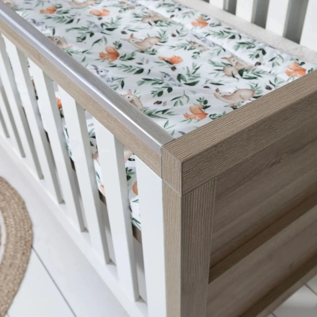 Close-up of light oak cot bed corner with white vertical rail and printed mattress cover