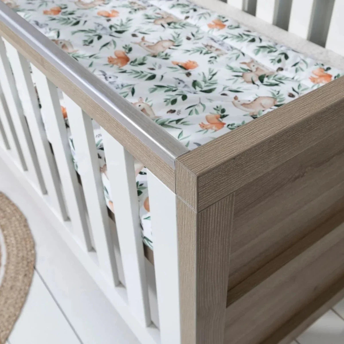 Close-up of light oak cot bed corner with white vertical rail and printed mattress cover