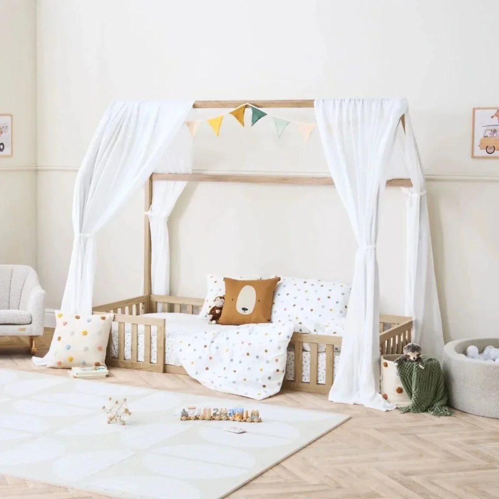 Oak house-style toddler floor bed with white curtains, bunting garland, and dotted bedding