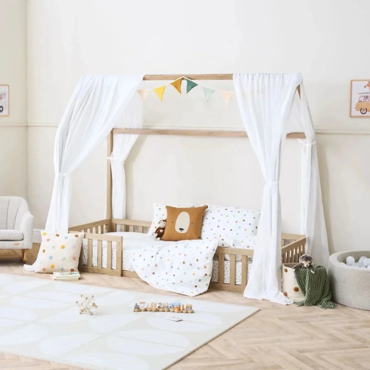 Oak house-style toddler floor bed with white curtains, bunting garland, and dotted bedding