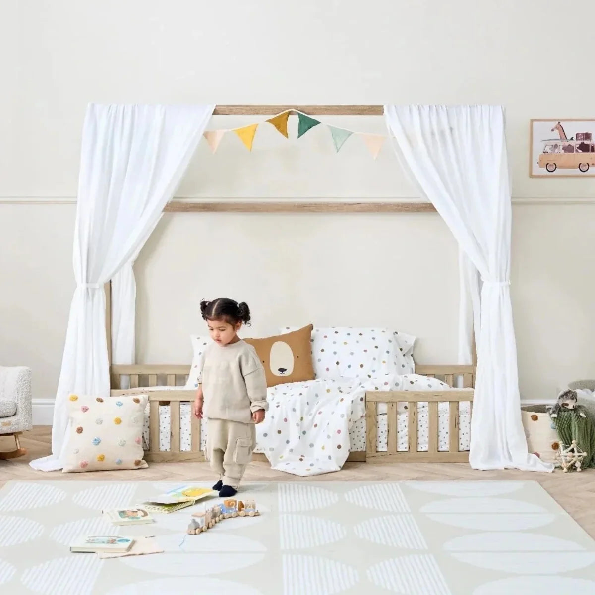 Oak house-style toddler floor bed with white curtains, dotted bedding, and toddler inside