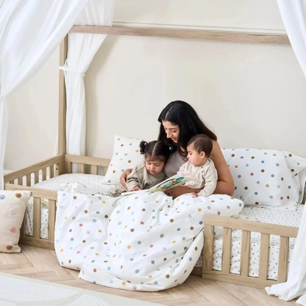 Oak toddler floor bed with white curtains and dotted bedding, children lying on mattress