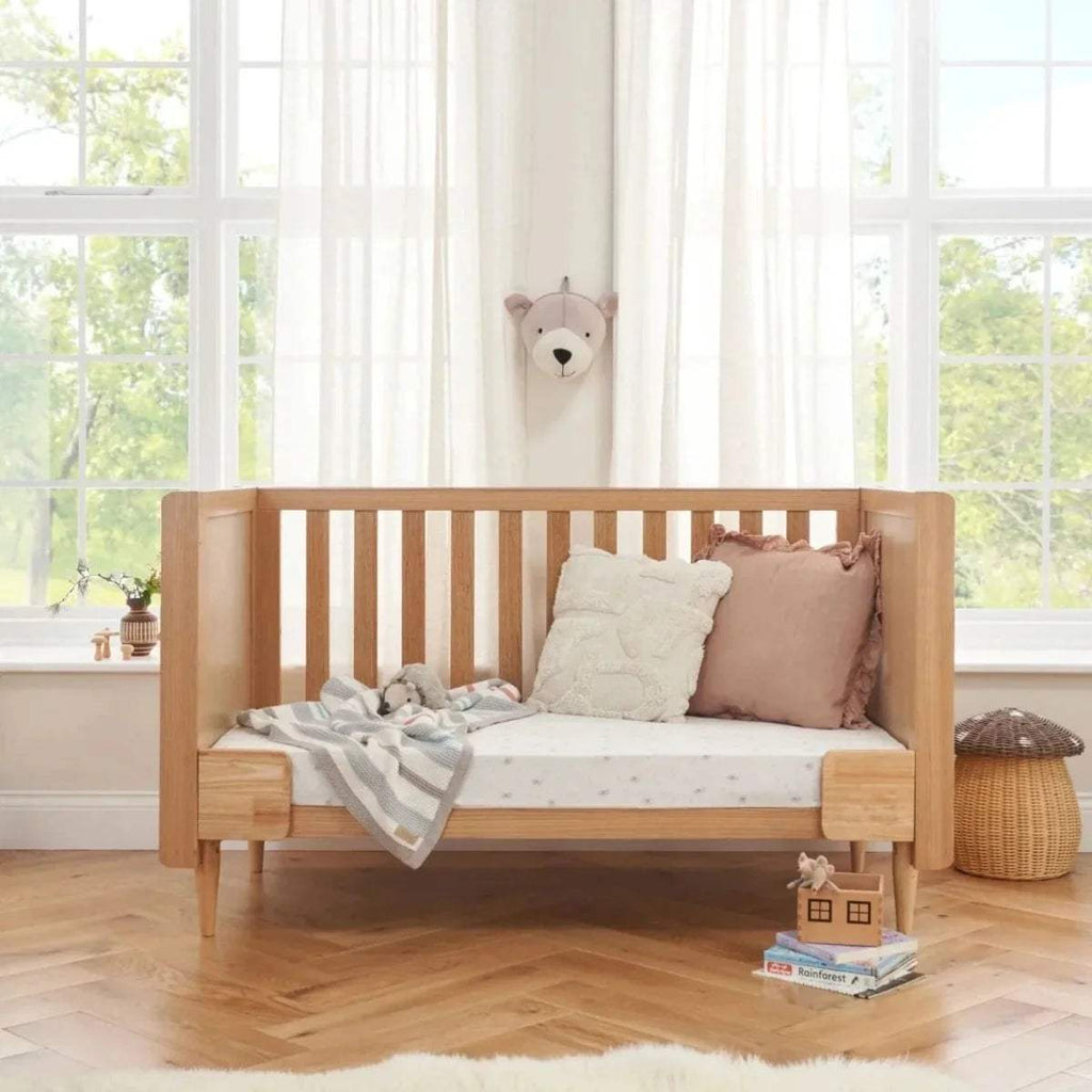 Light oak cot bed front view with vertical slat rails and cushioned bedding