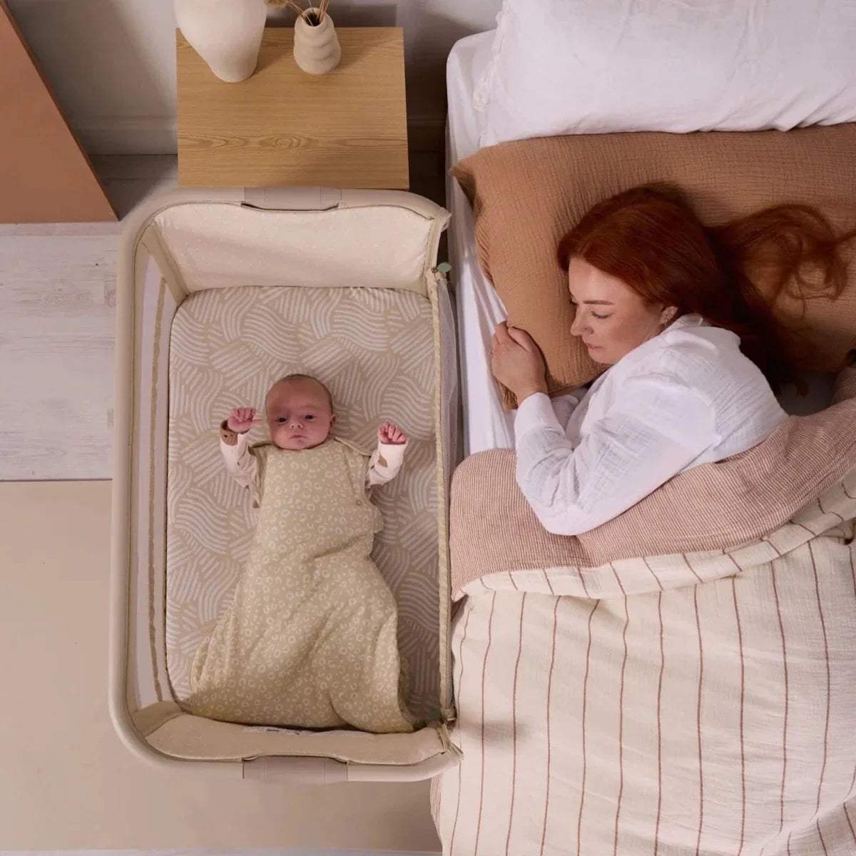 Sand bedside crib with baby lying on patterned pad, side rail raised