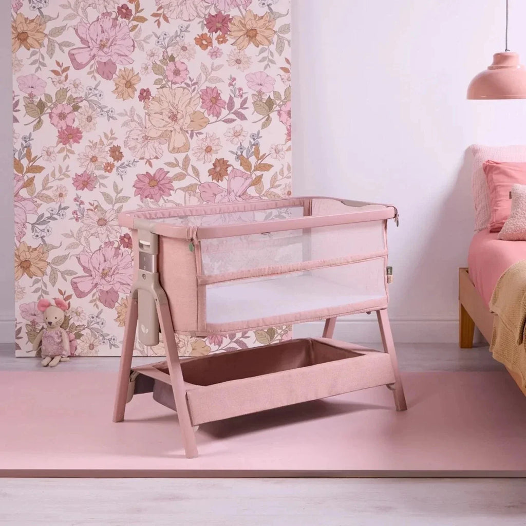 Blush bedside crib with open side and padded mattress in a mesh frame.