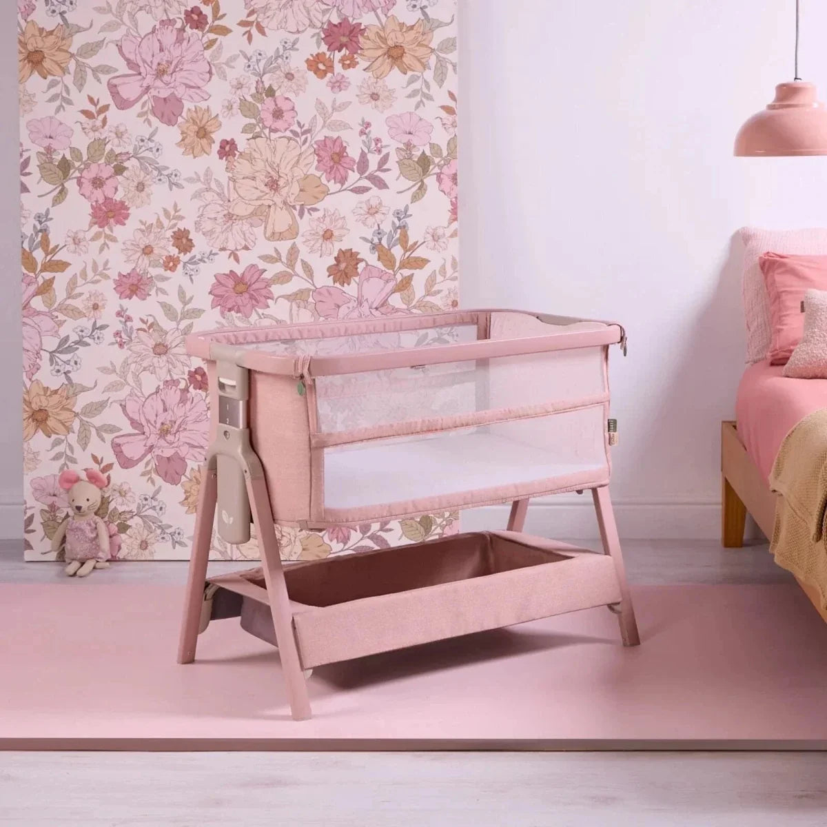 Blush bedside crib with open side and padded mattress in a mesh frame.