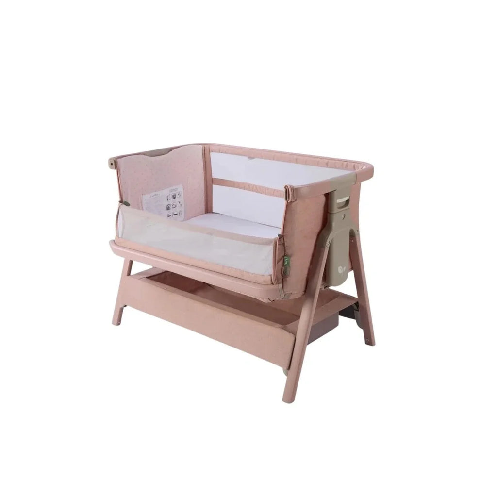 Blush bedside crib with mesh sides and wood frame, angled view.