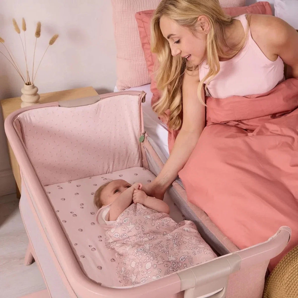 Blush bedside crib with baby in a patterned sleep sack on the padded mattress.