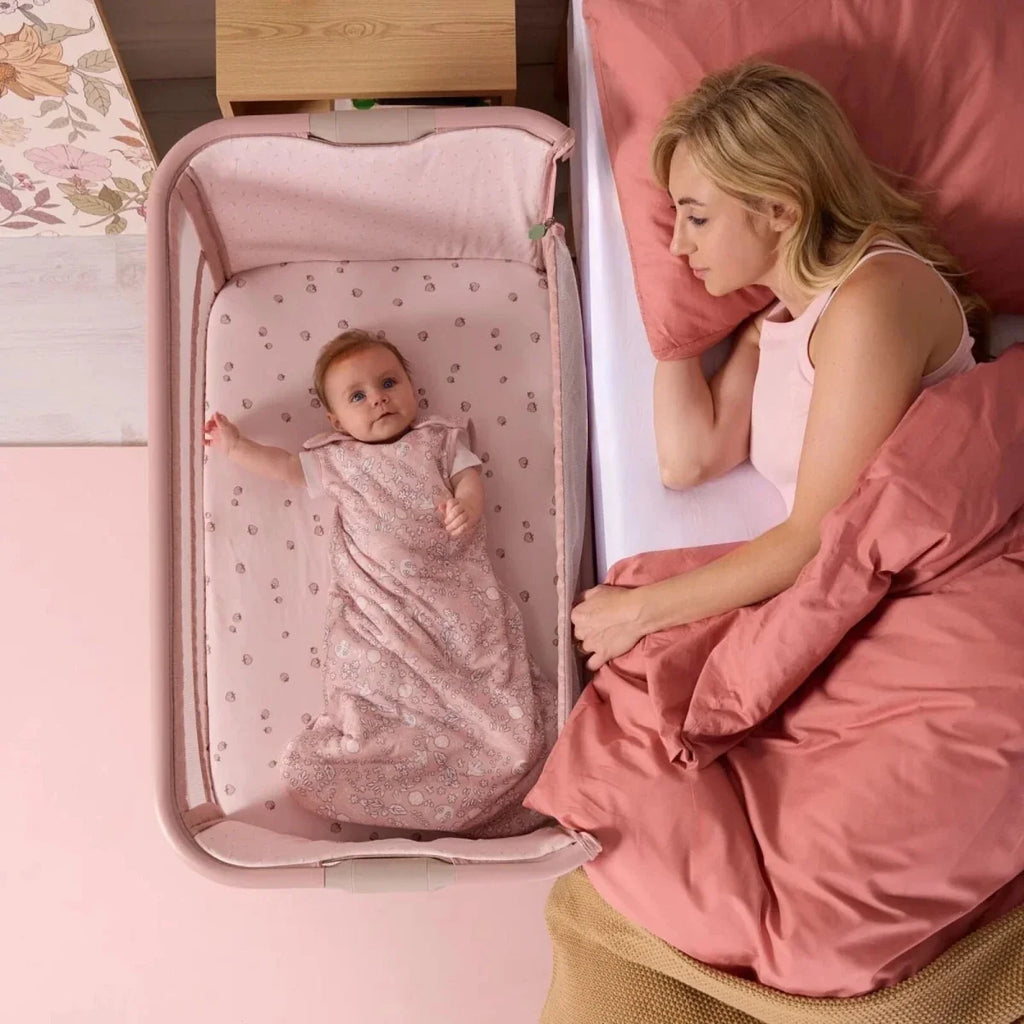 Blush bedside crib with baby lying on a padded mattress in the mesh frame.