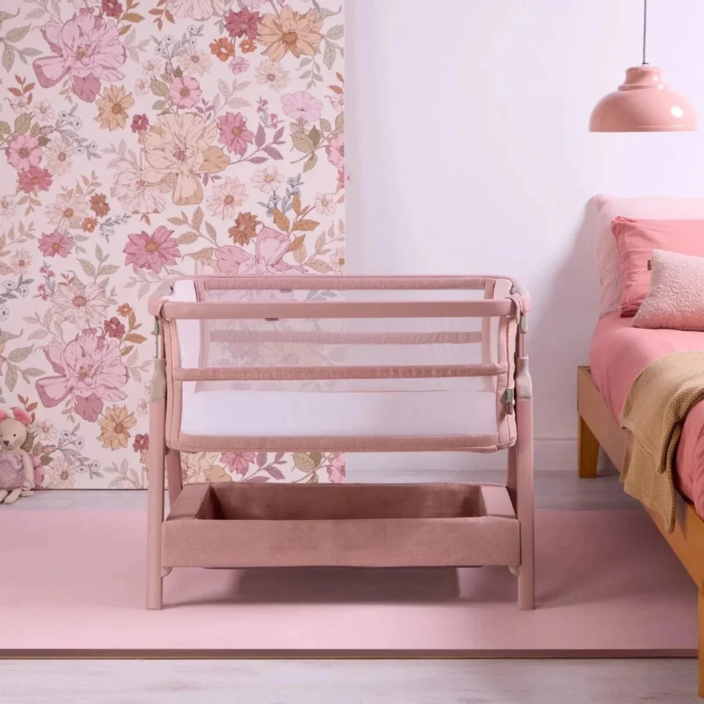 Blush bedside crib with mesh mattress area and front storage drawer.