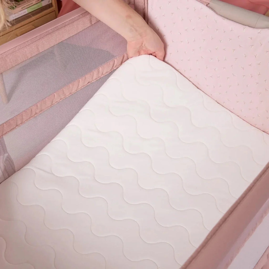 Blush bedside crib with white quilted mattress pad and raised side rails