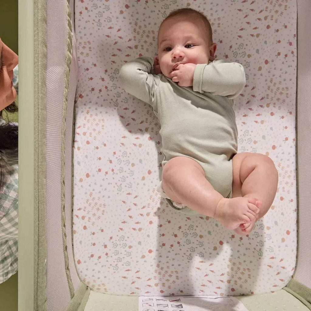 Baby in eucalyptus green bedside crib on patterned mattress with side rail visible