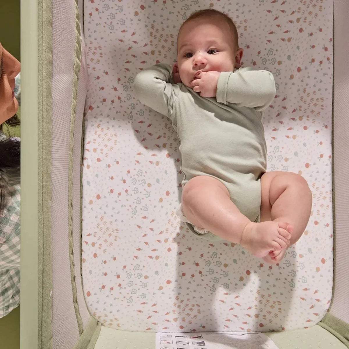 Baby in eucalyptus green bedside crib on patterned mattress with side rail visible