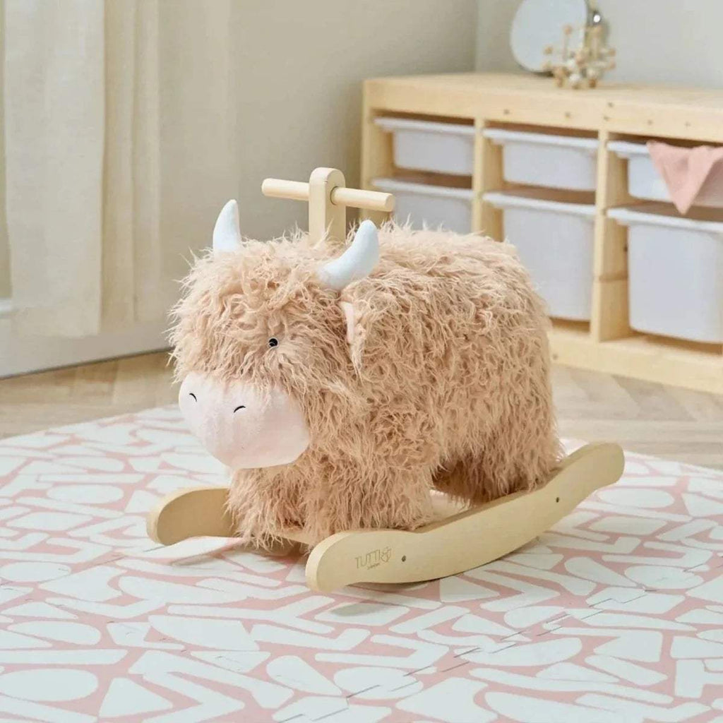 Highland cow rocking toy with beige plush body, white muzzle, and wooden rocker base.