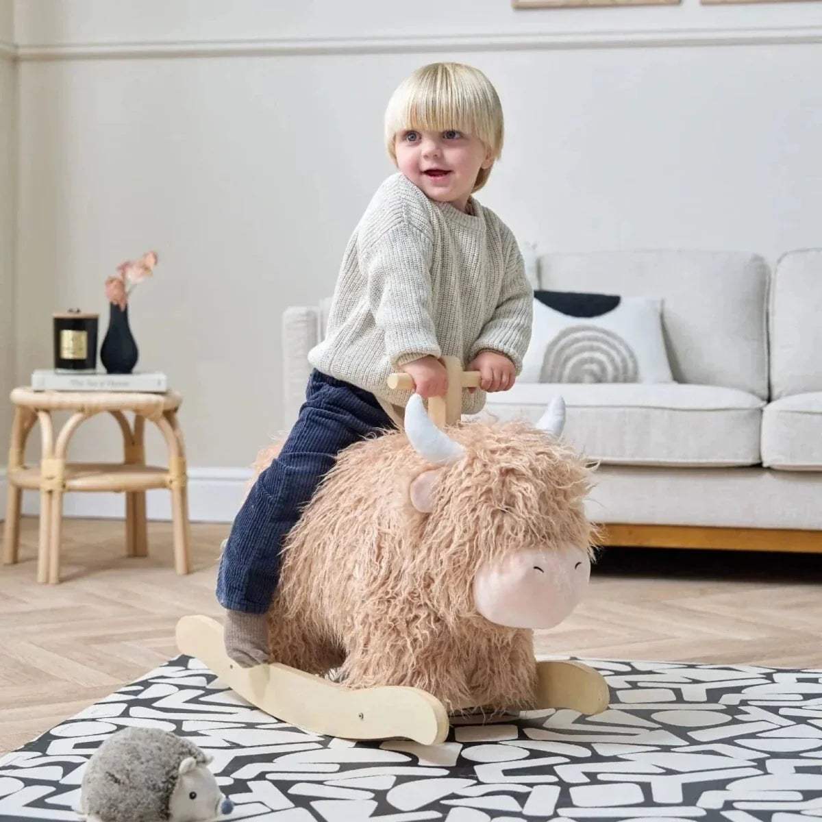 Highland cow rocking toy with beige plush fur and wooden rocker base, child in grey sweater.