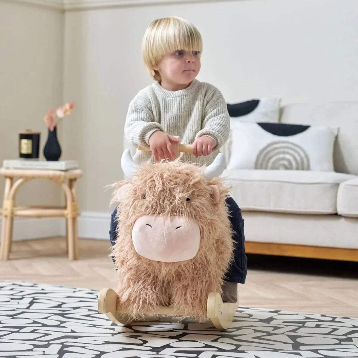 Highland cow rocking toy with beige plush face and wooden rocker base; child holding handles.