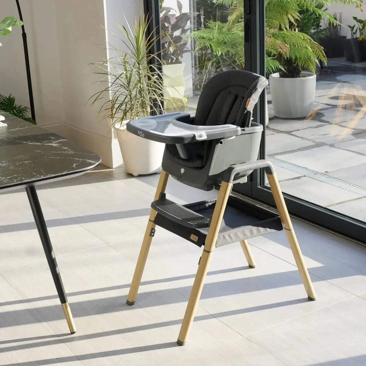 Grey highchair with tray and light oak legs, side view indoors on tiled floor.
