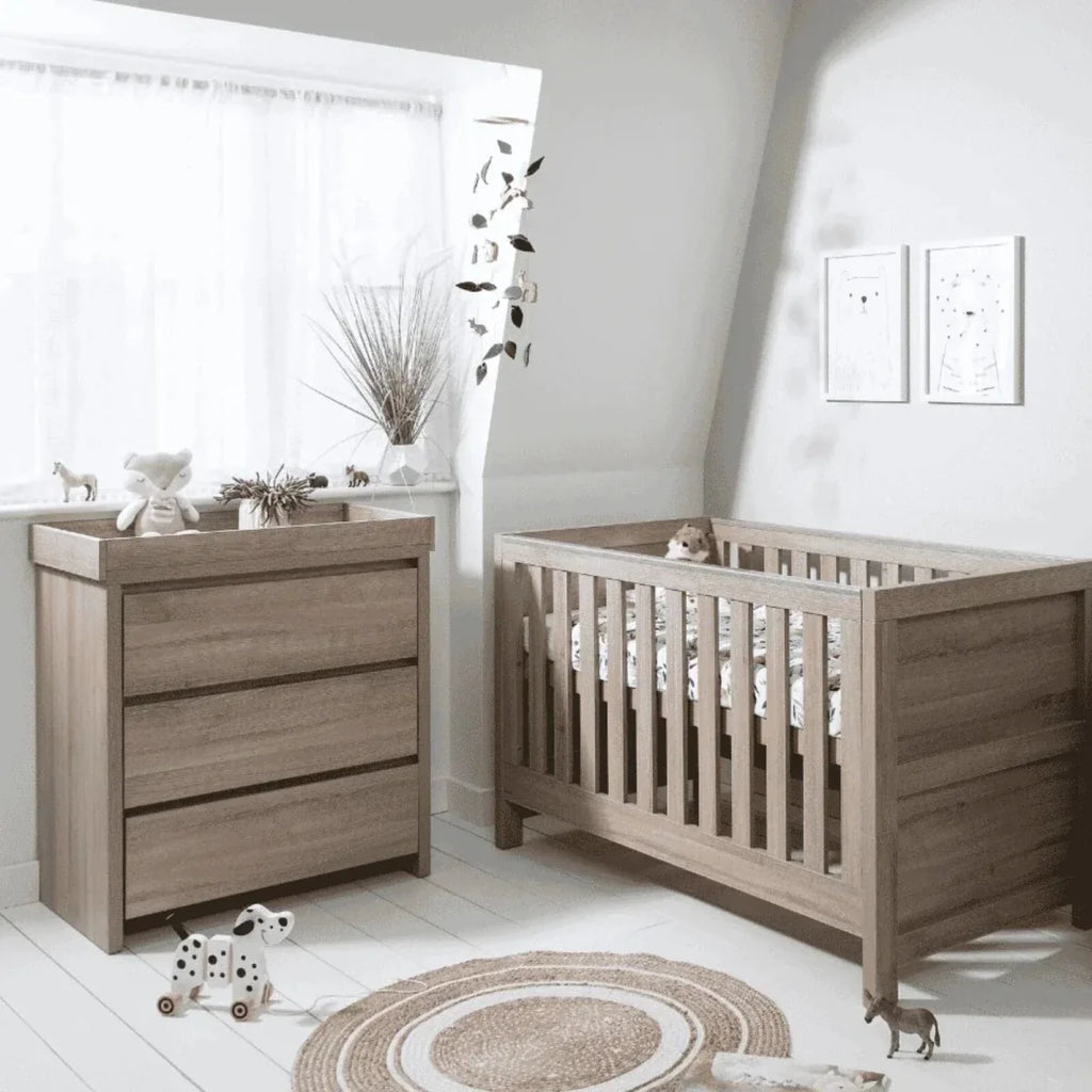 Light oak cot bed with vertical slats beside matching dresser with three drawer fronts.
