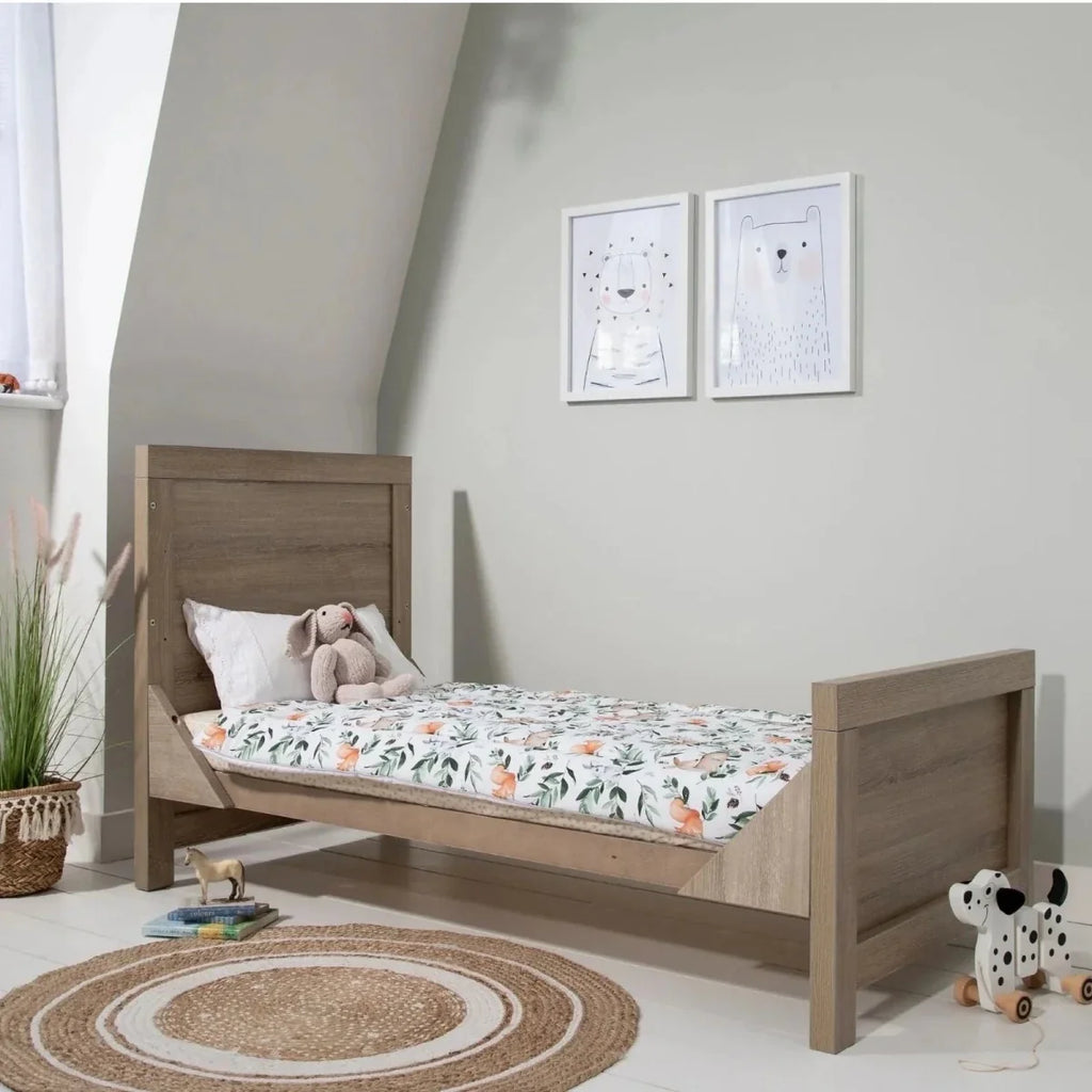 Oak cot bed with white patterned bedding and pillows, set in a nursery corner.
